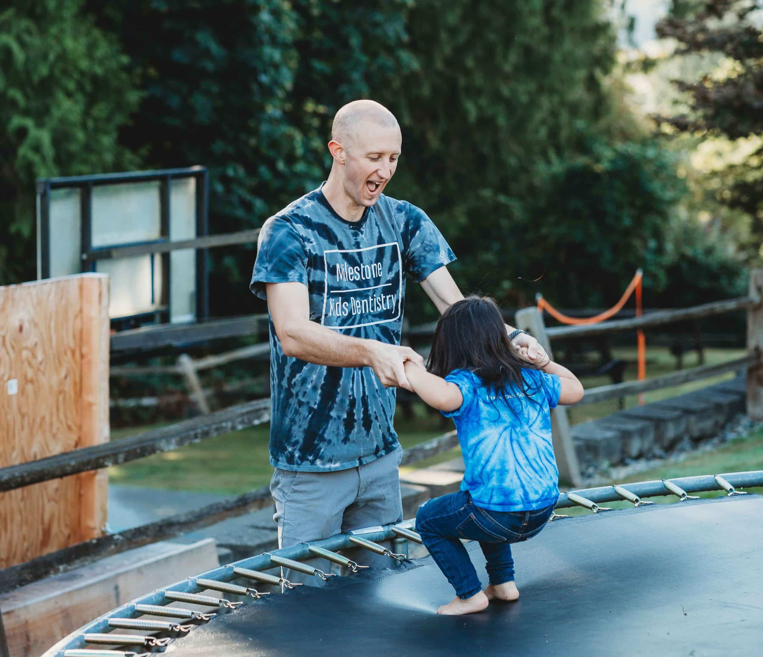 A man holding the hands of a young girl who is jumping on a trampoline.
