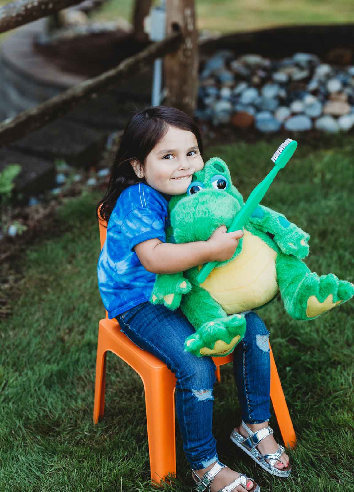 A girl in a tie-dye shirt, holding a large green toothbrush and hugging a plush dinosaur.