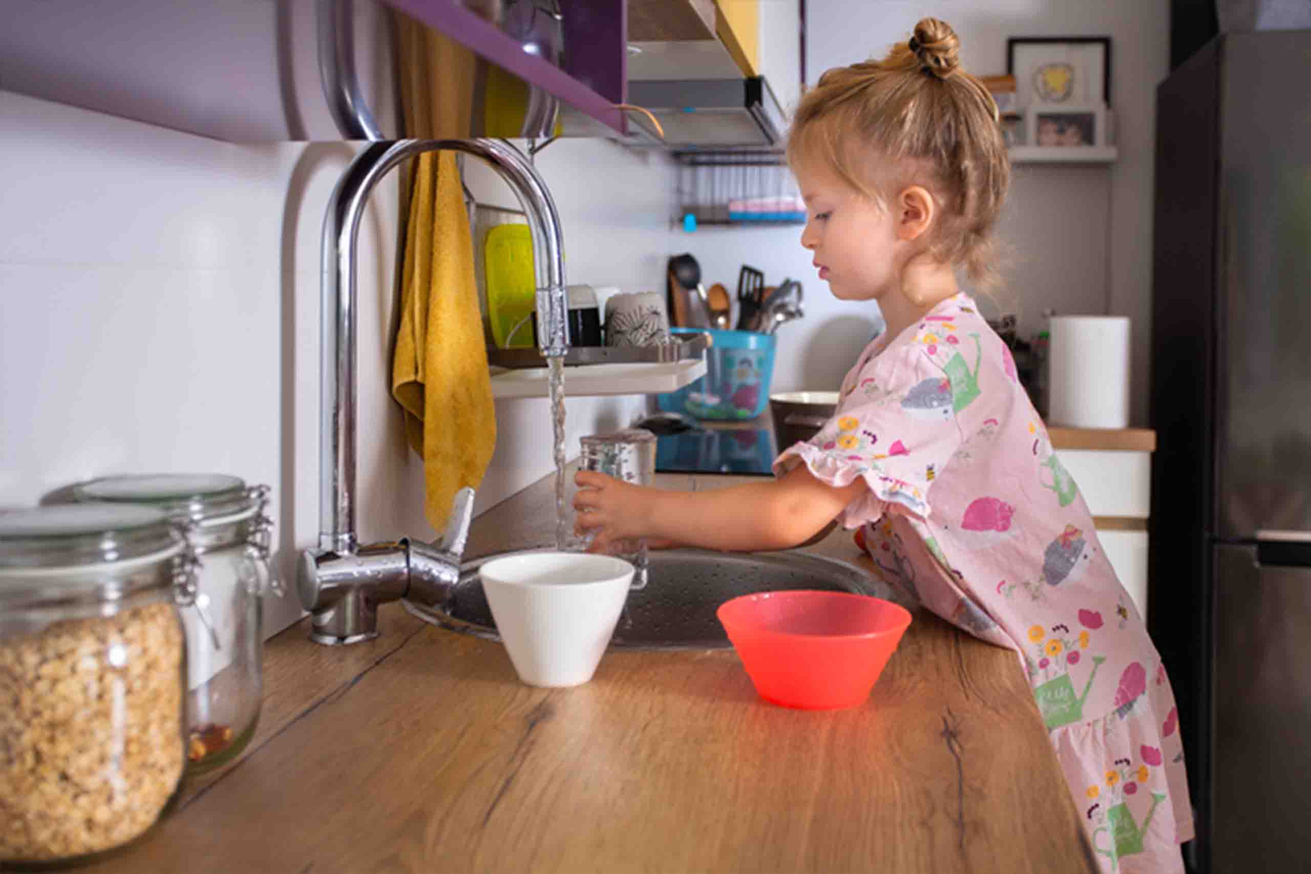 A girl in a pink dress filling a cup of tap water in her kitchen.