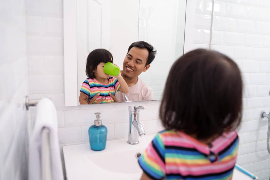 A young girl rinsing with a green cup of mouthwash while her dad supervises.
