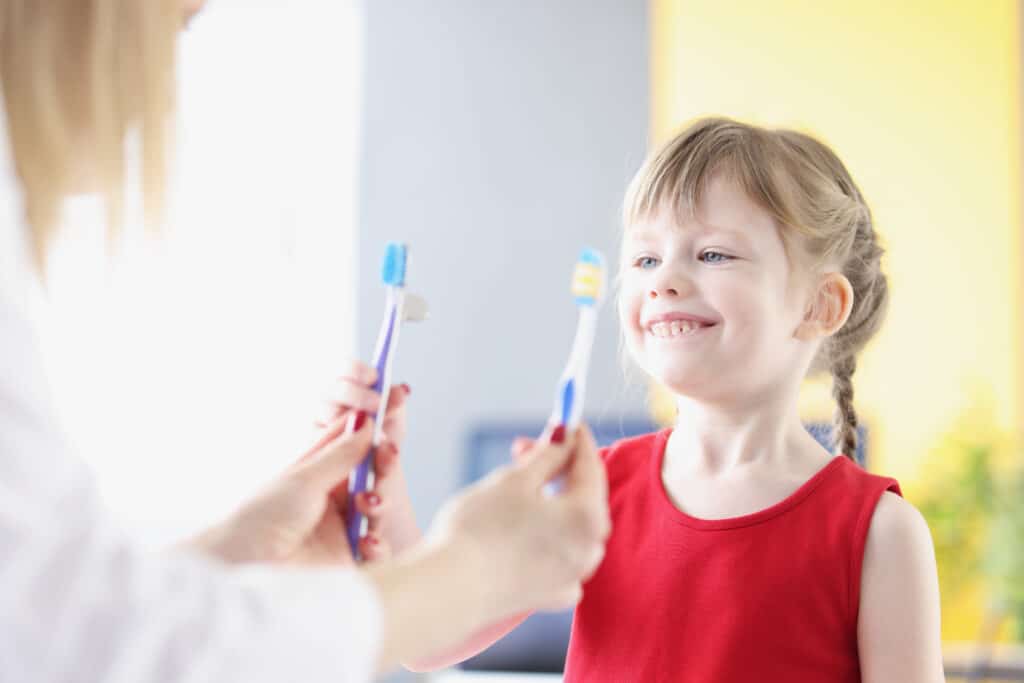 A young girl in a red shirt choosing between 2 toothbrushes that a dentist is holding.